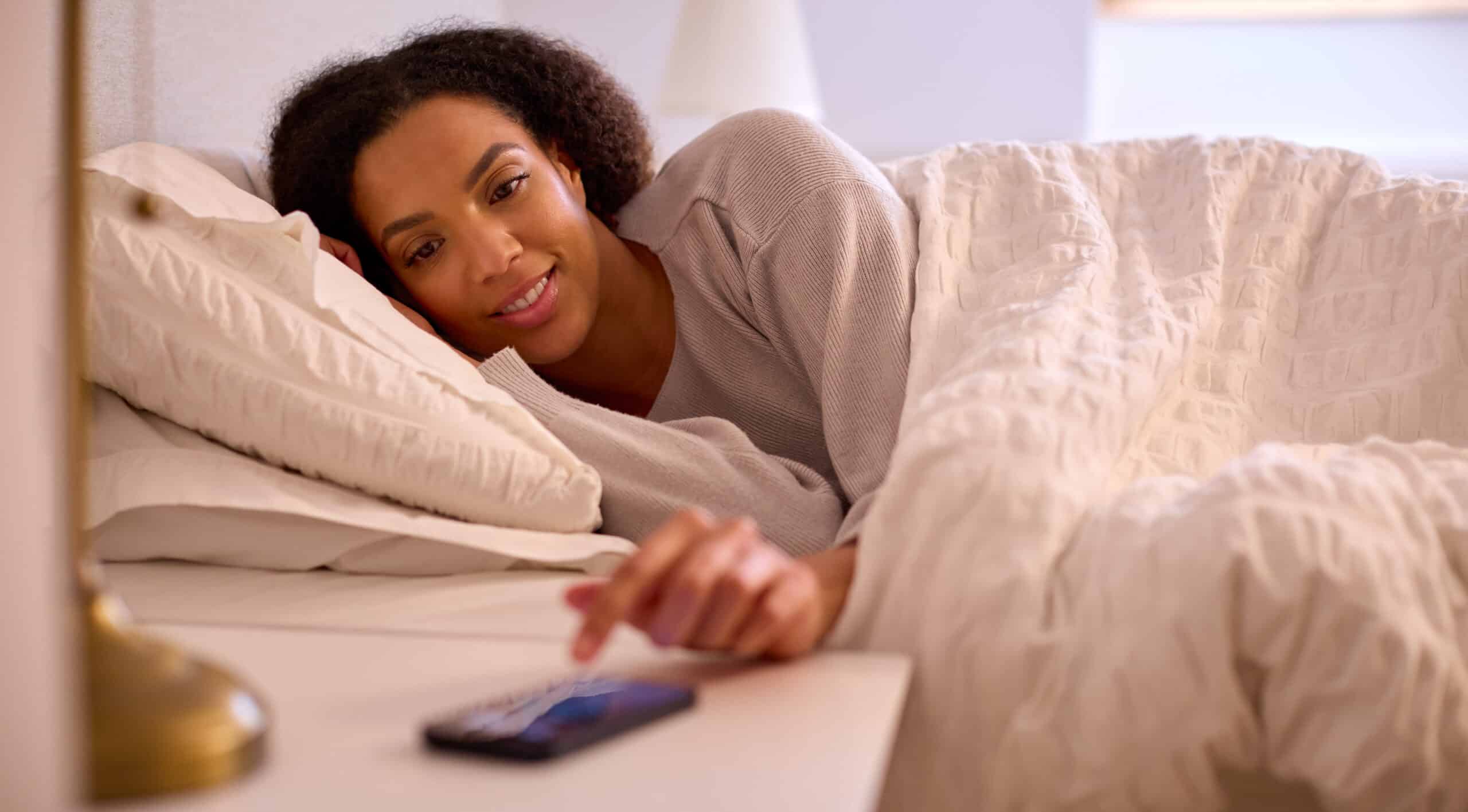 woman at home in bed turning off alarm on mobile phone