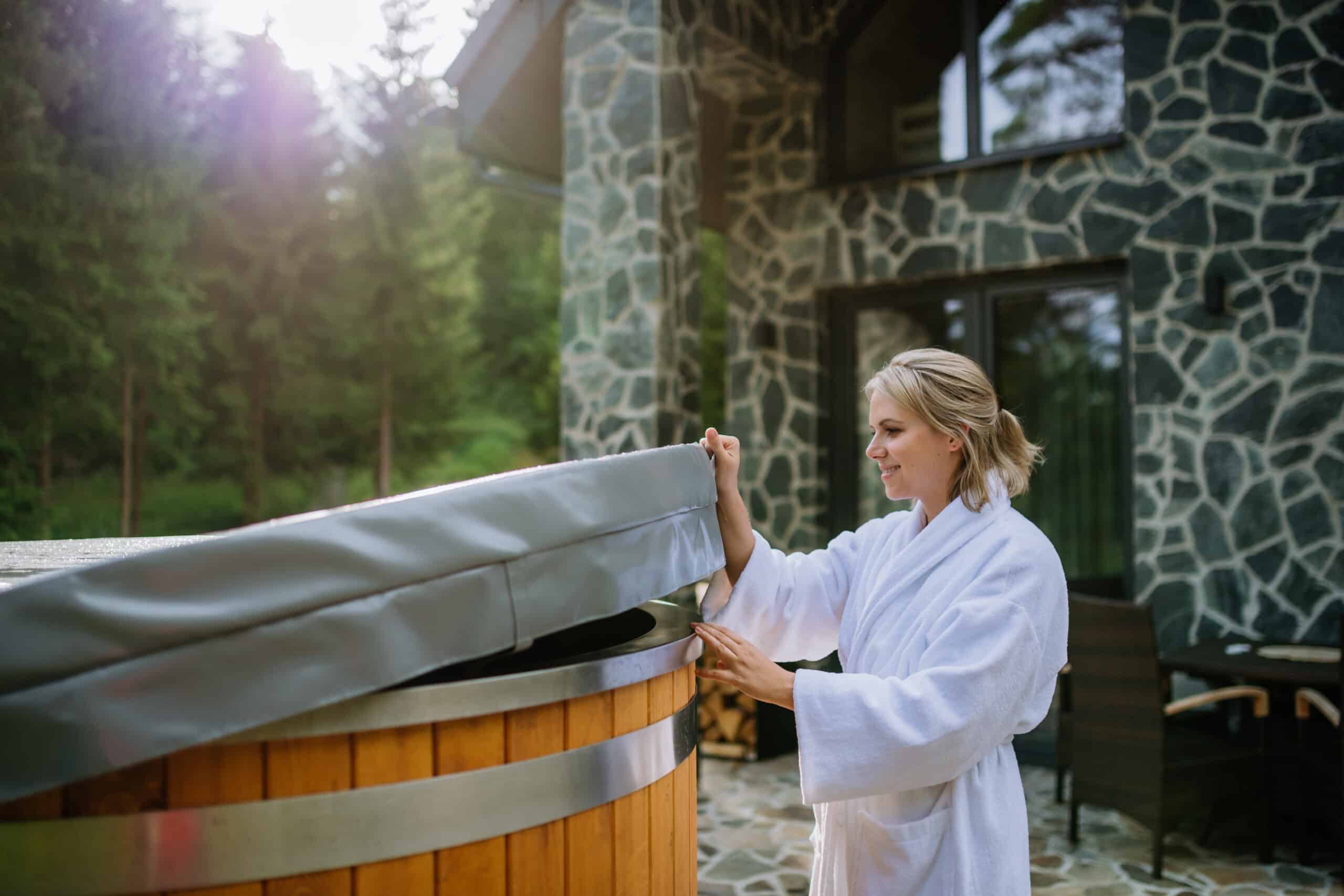 woman in bathrobe opening lid of hot tub, checking temperature, ready for home spa procedure in hot tub outdoors. wellness, body care, hygiene concept.